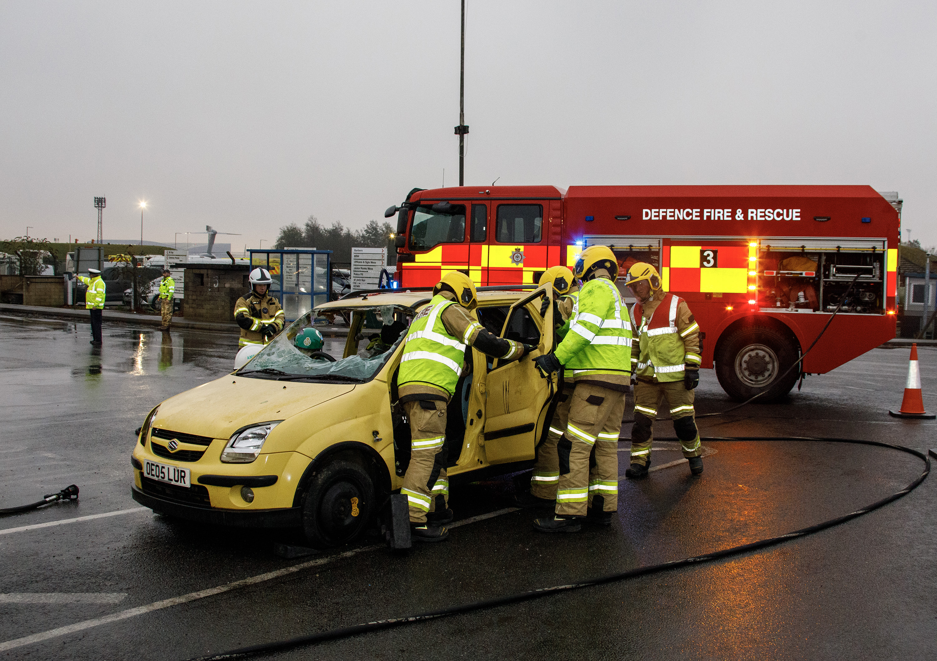 Photo - Image shows RAF police managing and directing traffic flow and a Defence Fire and rescue vehicle in the background, with fire personnel next to the car, on the passenger side, having opened the rear passenger door.
